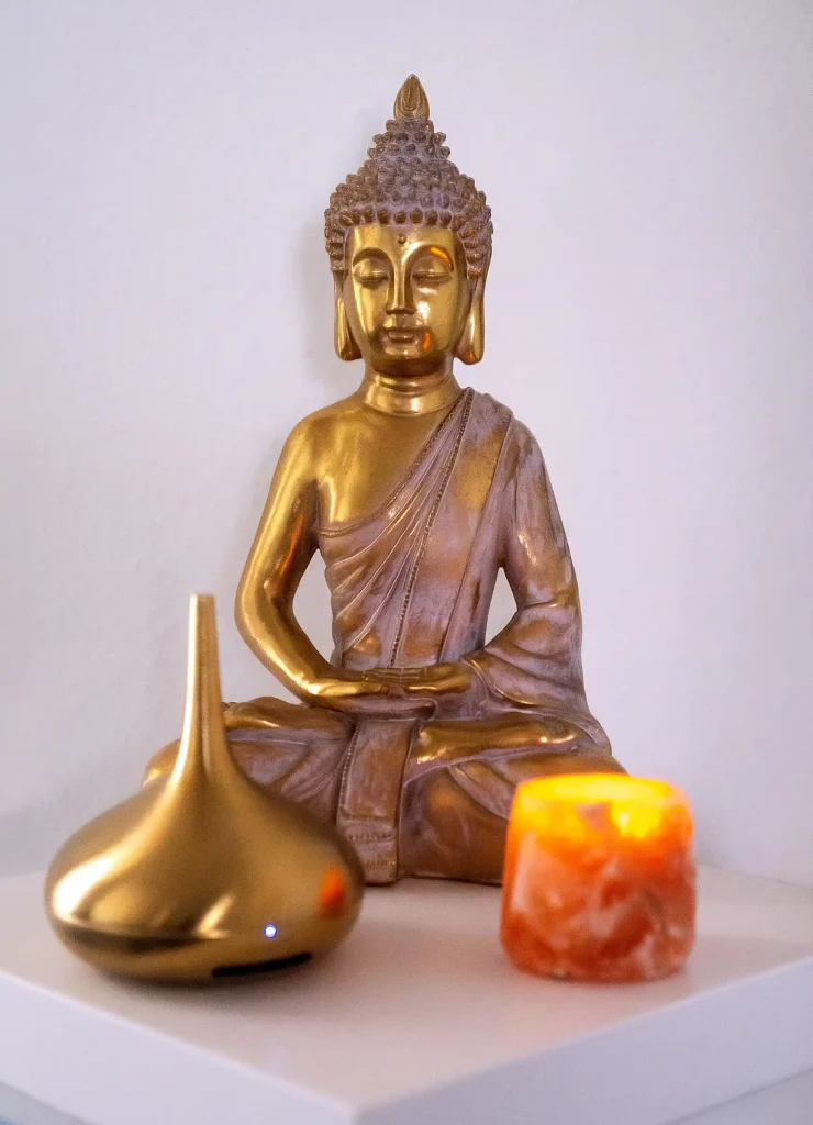 Goldglänzende Buddhastatue in Meditationshaltung, flankiert von einer funkelnden Salzleuchte und einer stilvollen goldenen Halterung für Räucherstäbchen.